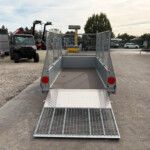 Open metal utility trailer with a grated ramp extended into a gravel lot, surrounded by heavy machinery and vehicles in an industrial yard.