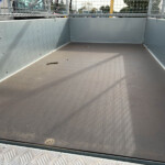Empty gray metal trailer bed with riveted side walls and a diamond-plate floor, parked in a lot behind a mesh railing.