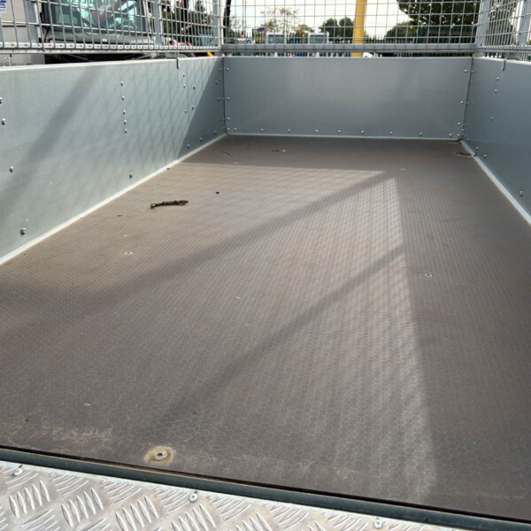 Empty gray metal trailer bed with riveted side walls and a diamond-plate floor, parked in a lot behind a mesh railing.