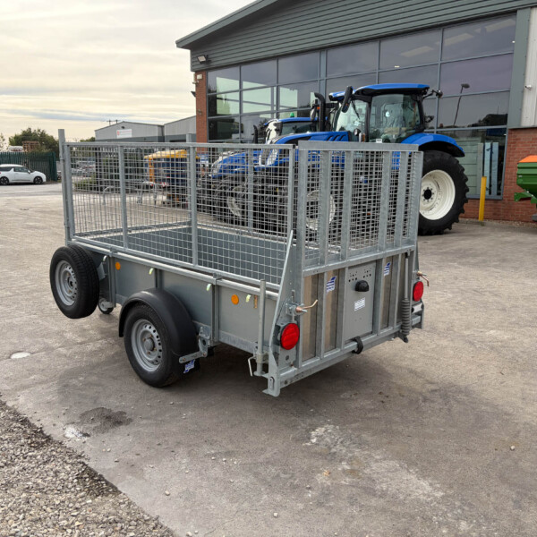 Metal open-sided utility trailer in the foreground with a blue tractor behind it in an industrial parking lot near a glass-fronted building.