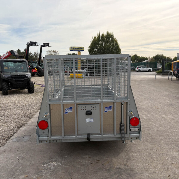 Rear view of a gray metal utility trailer in a equipment lot, with farm machinery and a New Holland sign in the background, under a cloudy sky.