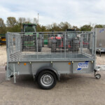 Grey Ifor Williams trailer with metal mesh sides in a gravel lot, a green tractor behind it and trees in the background.