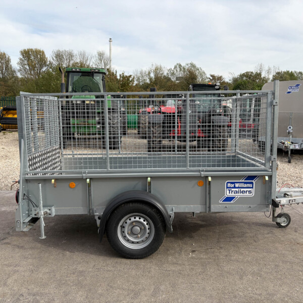 Grey Ifor Williams trailer with metal mesh sides in a gravel lot, a green tractor behind it and trees in the background.