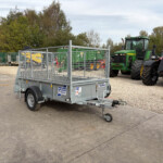 A metal flatbed trailer with mesh sides parked on a gravel lot, with large green tractors and equipment in the background.