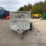Front view of a galvanized utility trailer with mesh sides in a gravel lot, with industrial equipment and a green dumpster in the background.