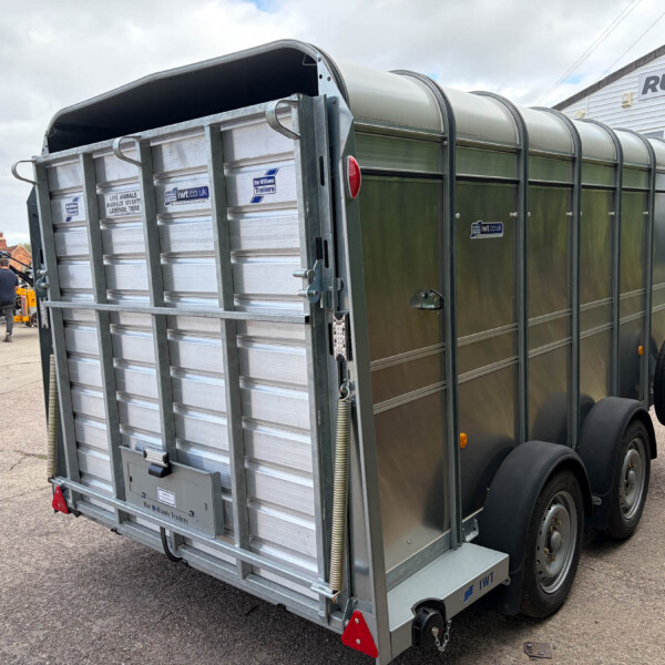 Rear view of a large metal livestock trailer with curved roof panels in a dealership lot. Onlookers and other trailers are in the background.