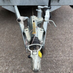 Close-up of a silver trailer hitch and jack assembly with a frayed protective cover on a gravel surface.
