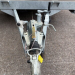 Close-up of a trailer hitch showing the coupler, tongue jack, safety chains, and wiring on a gravel surface.