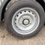 Close-up of a tire mounted on a silver steel trailer wheel with five lug nuts and a visible valve stem, resting on a gravel surface under a fender.