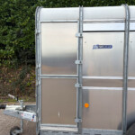 Front corner of a silver metal trailer with latches and hinges, outdoors beside a dense green hedge and via paved ground edge