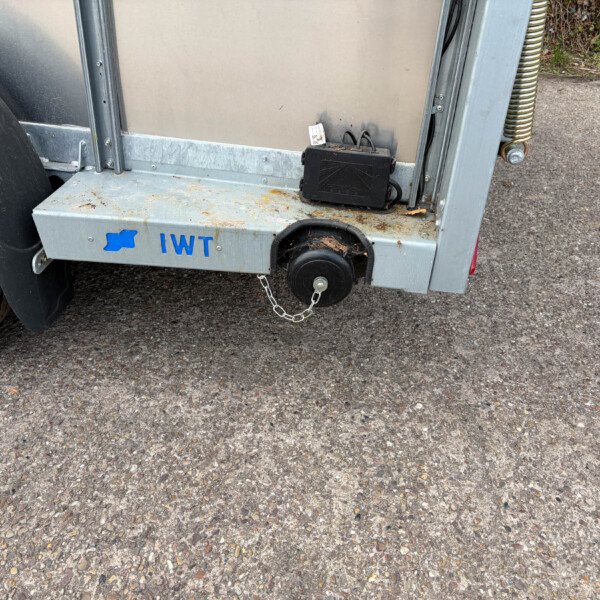 Back end of a metal trailer with a blue IWT logo, showing a hitch, spring, and a rusty, dirty frame near a gravel surface.