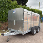 Silver metal trailer with curved roof and riveted panels, parked on a street beside a tall green hedge (Ifor Williams Trailers logo visible).