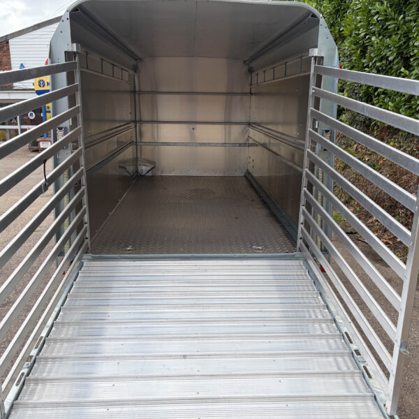 Empty galvanized metal trailer with side rails and diamond-plate floor, open at rear outdoors by a hedge and store frontage
