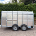 Silver aluminum trailer with rounded top and dual axles parked on a paved surface, hedge in the background.