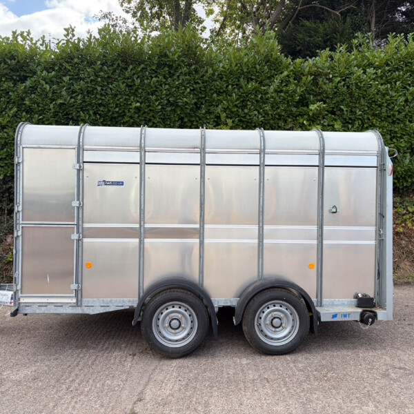 Silver aluminum trailer with rounded top and dual axles parked on a paved surface, hedge in the background.