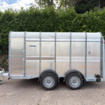 Silver metal trailer with a rounded top parked on a paved lot, dense green hedges behind it. Minors: two wheels visible and hitch at the front left.