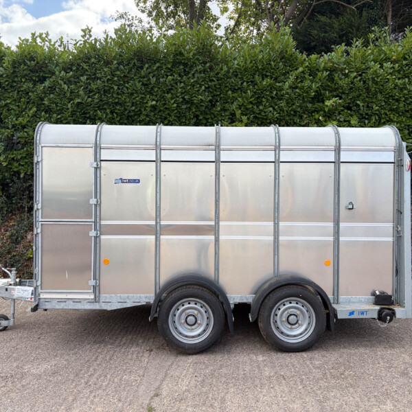 Silver metal trailer with a rounded top parked on a paved lot, dense green hedges behind it. Minors: two wheels visible and hitch at the front left.