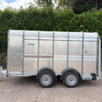 Silver enclosed utility trailer with two axles parked on a paved surface in front of a tall green hedge.