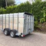 Silver metal enclosed trailer with curved roof panels parked on an asphalt lot, backed by a tall green hedge; a red car is visible on the left edge.