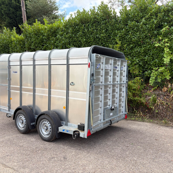 Silver metal enclosed trailer with curved roof panels parked on an asphalt lot, backed by a tall green hedge; a red car is visible on the left edge.