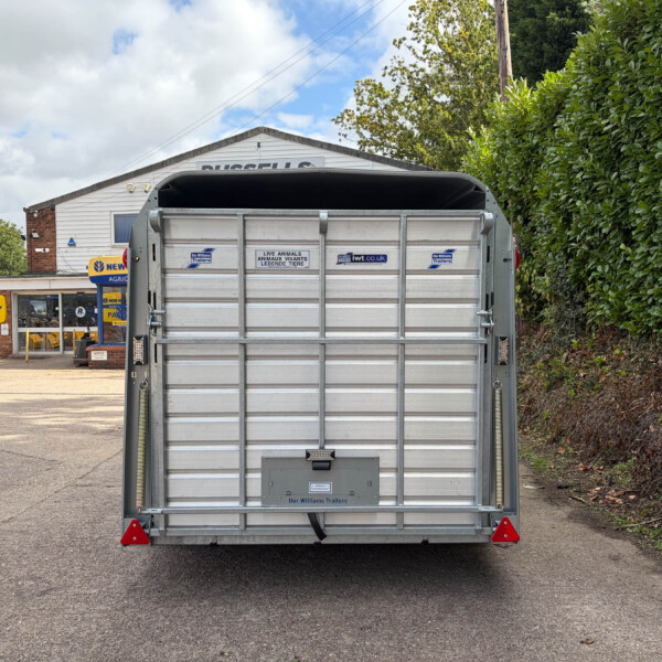 Rear view of a silver livestock trailer with closed gates, parked in a service yard near a building and hedge in daytime.
