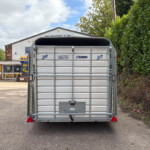 Rear view of a metal trailer parked in a lot, with a building and signage in the background and dense hedges to the right.
