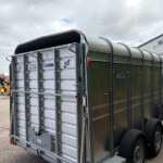 Rear of a metal livestock trailer with a curved roof and twin axles parked in a dealership lot, with a Russell building in the background.