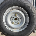 Close-up of a black tire mounted on a silver steel wheel with five lug nuts; outdoor equipment visible in the background.