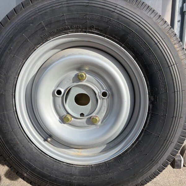 Close-up of a black tire mounted on a silver steel wheel with five lug nuts; outdoor equipment visible in the background.