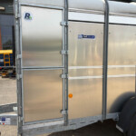 Side view of a silver metal trailer with riveted panels and branding stickers (iwtt.co.uk, Russell). | Close-up of a large trailer door with latches and a wheel, logos visible.
