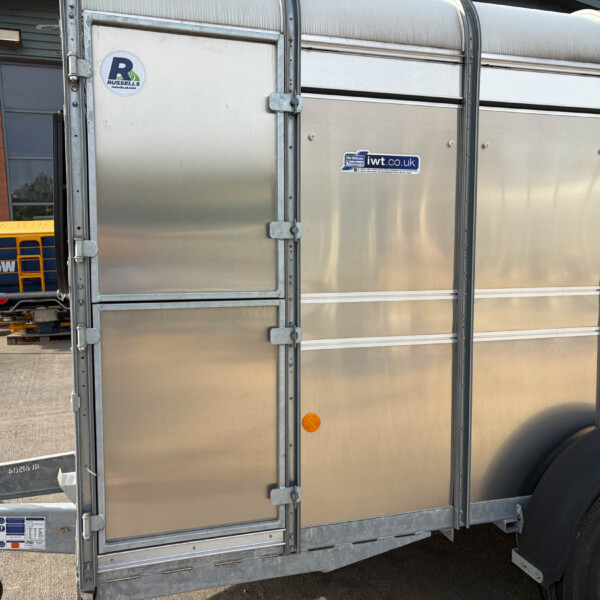 Side view of a silver metal trailer with riveted panels and branding stickers (iwtt.co.uk, Russell). | Close-up of a large trailer door with latches and a wheel, logos visible.