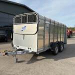 Silver livestock trailer with curved vented front, hitched outside a building with equipment in the background