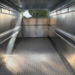 Empty aluminum cargo trailer interior with diamond-plate floor and mesh rear gate exposing trees outside.