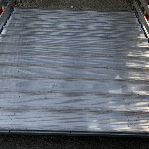 Top-down view of ribbed aluminum stairs on a loading platform with side rails and a dark ground below.