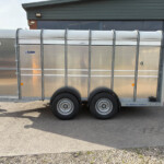 Long silver livestock trailer with curved roof panels parked in an industrial lot, attached to a blue tractor on the right.