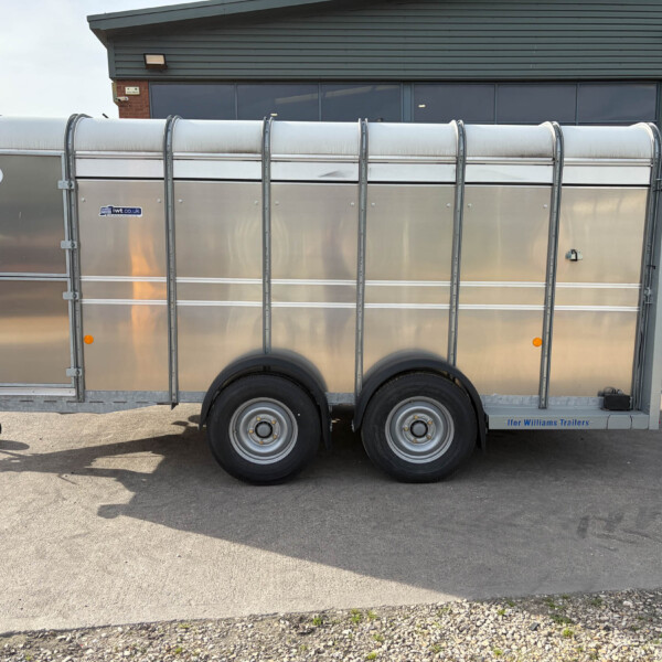 Long silver livestock trailer with curved roof panels parked in an industrial lot, attached to a blue tractor on the right.