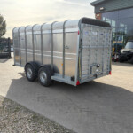 Metal silver livestock trailer with dual axles parked in a lot near a glass-front building and a tractor nearby.