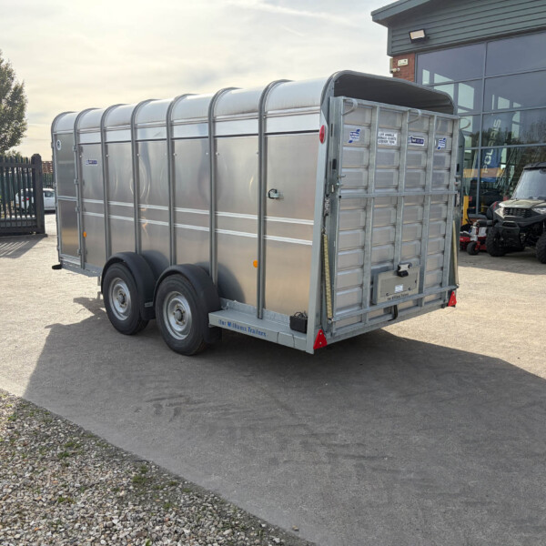 Metal silver livestock trailer with dual axles parked in a lot near a glass-front building and a tractor nearby.