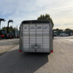 Rear view of a silver metal trailer in a gravel lot, with red agricultural tractors lined up on the left and a fence in the background.