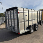 Rear view of a large silver livestock trailer with curved roof and dual-axle wheels, parked on asphalt with a New Holland Agriculture sign in the background.