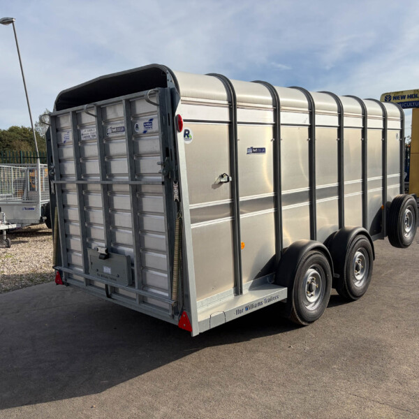 Rear view of a large silver livestock trailer with curved roof and dual-axle wheels, parked on asphalt with a New Holland Agriculture sign in the background.