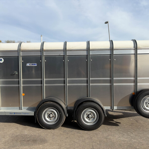 Side view of a long silver livestock trailer with multiple wheels parked on a paved lot against a blue sky.