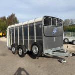 Three-axle gray livestock trailer with rounded roof panels and metal ventilation grid, parked in an outdoor yard.