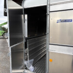 Open metal trailer with dark interior and diamond-plate floor; a lime-green construction loader is visible outside left.