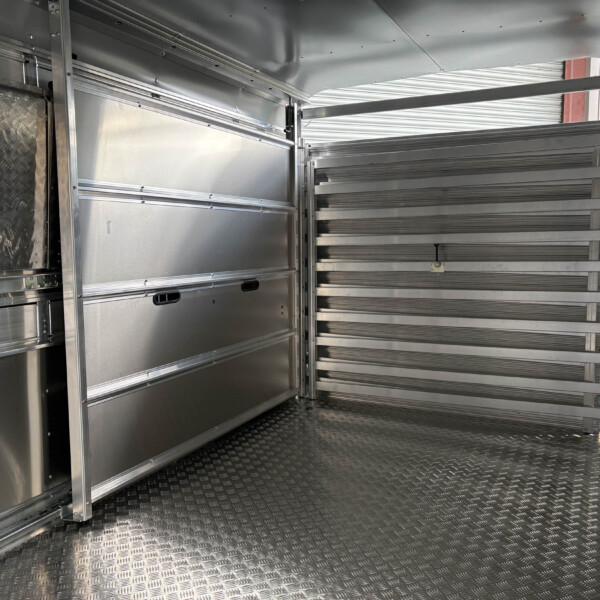 Interior of a metal trailer showing aluminum walls, ribbed panels, and diamond-plate floor with a stair-step rack on the right side (empty loading area).