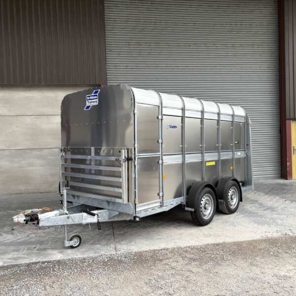 Silver aluminum trailer with rounded top rails parked outside a corrugated metal warehouse, hitch at the front visible.”