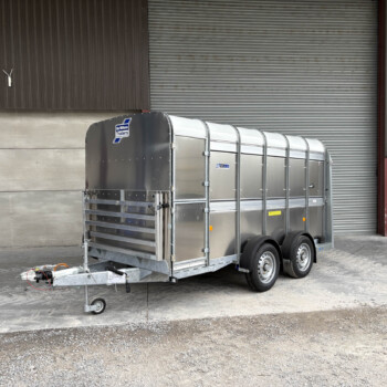 A metal livestock trailer hitched to a towing arm in front of a brown corrugated wall and a large gray roll-up door at an industrial site.