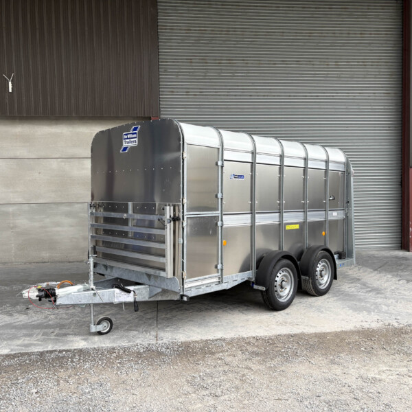 A metal livestock trailer hitched to a towing arm in front of a brown corrugated wall and a large gray roll-up door at an industrial site.