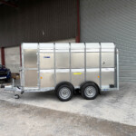 Silver aluminum enclosed trailer attached to a blue pickup under a large metal warehouse.