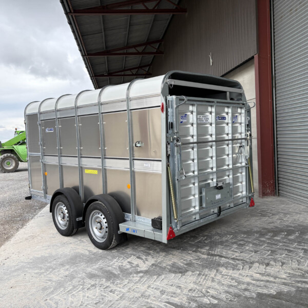 Silver livestock trailer parked beside a large warehouse with a corrugated metal roll-up door; farm equipment in the background on the left side of the image.